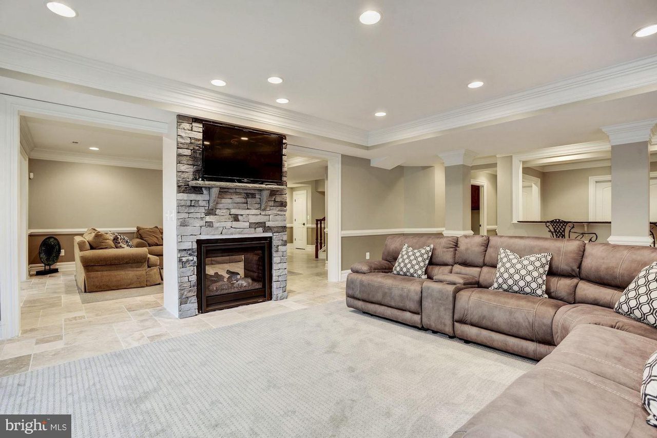 Spacious living room with a large sectional sofa, stone fireplace, wall-mounted TV, and recessed lighting. A separate room with another couch is visible through an open doorway.
