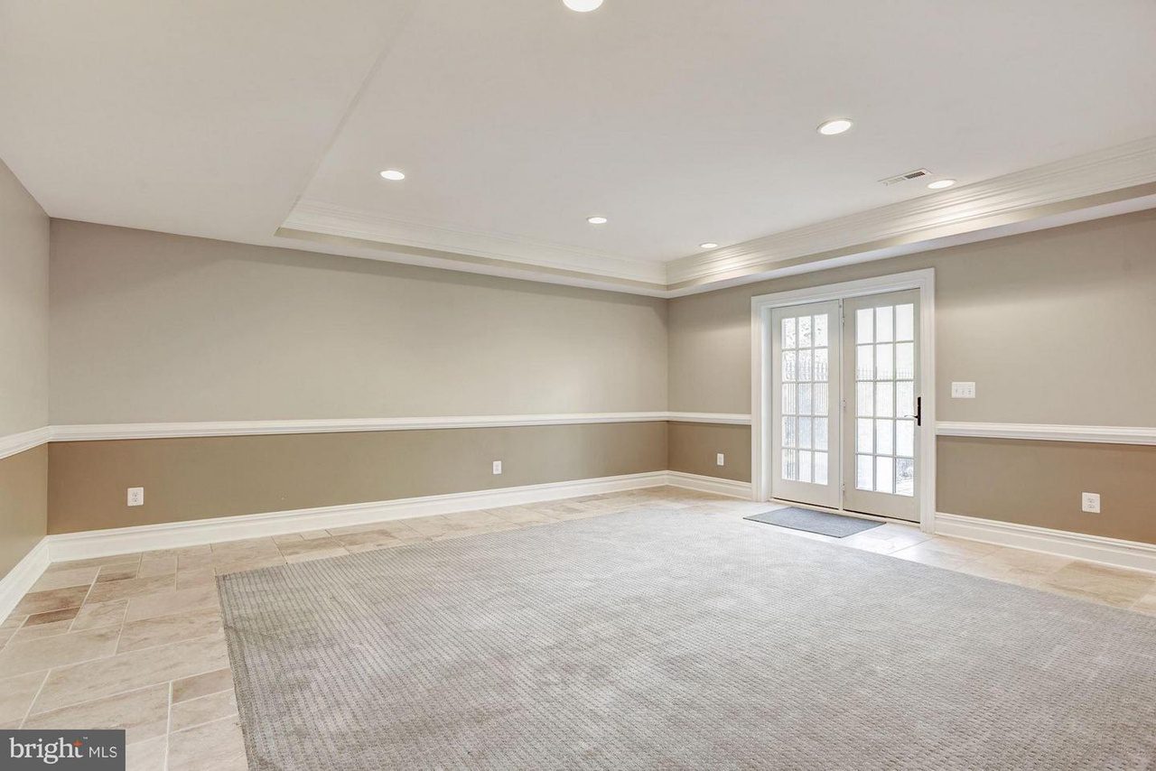 Empty room with beige walls, recessed ceiling lights, tiled floor, and a large gray rug. French doors on the right open to the outside.