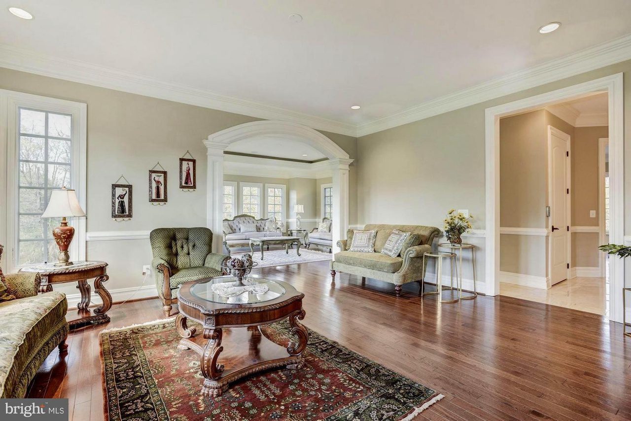 Spacious living room with hardwood floors, green sofas, a wooden coffee table, vintage rug, and arched doorway leading to a bright adjacent room. Walls are light-colored with decorative moldings.