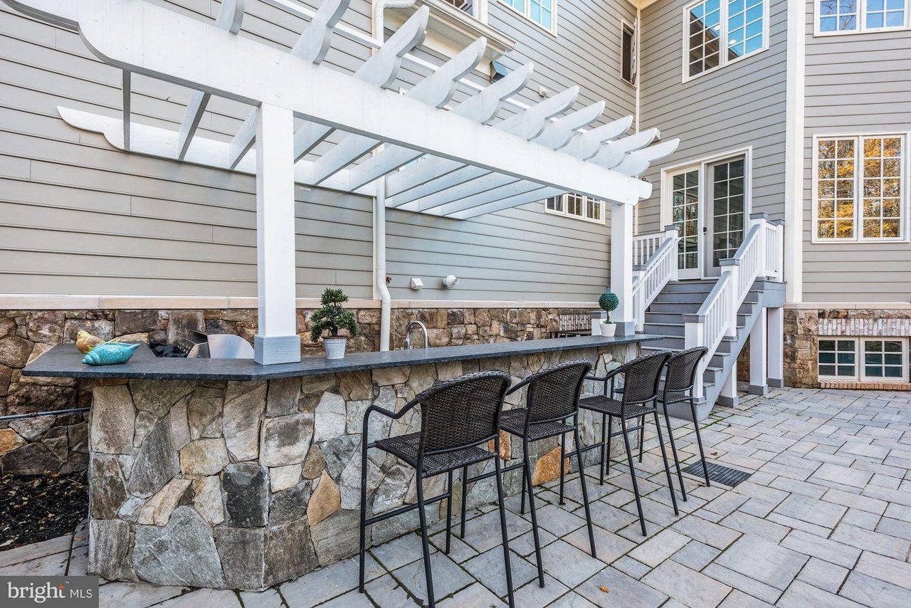 Outdoor patio with a stone bar beneath a white pergola. Four black barstools are lined up along the bar. Stairs lead up to a house with beige siding and several windows.