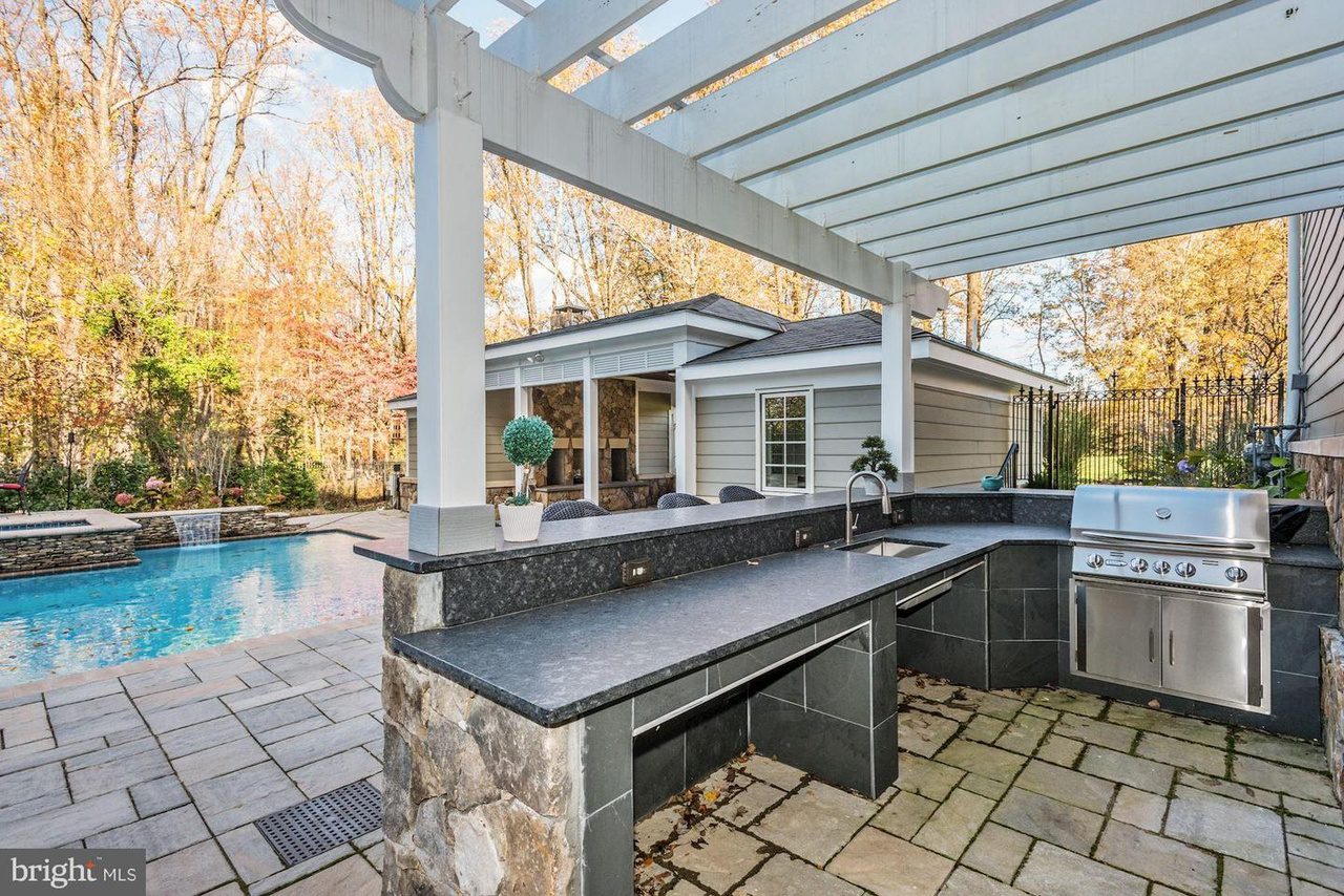 Outdoor kitchen with stone countertops, grill, and pergola next to a swimming pool surrounded by trees.