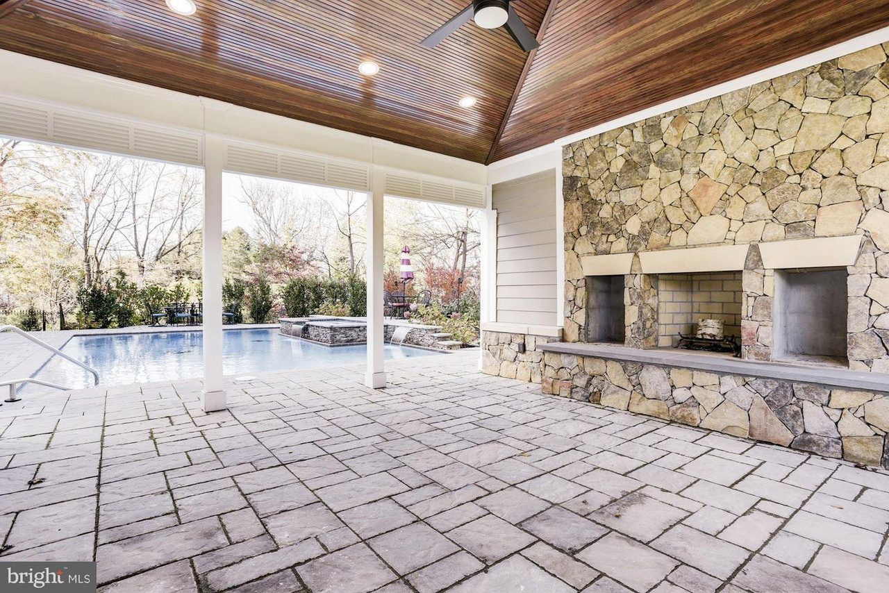 Outdoor patio with stone flooring and fireplace under a wooden ceiling, overlooking a pool with surrounding trees and a lighthouse structure in the background.