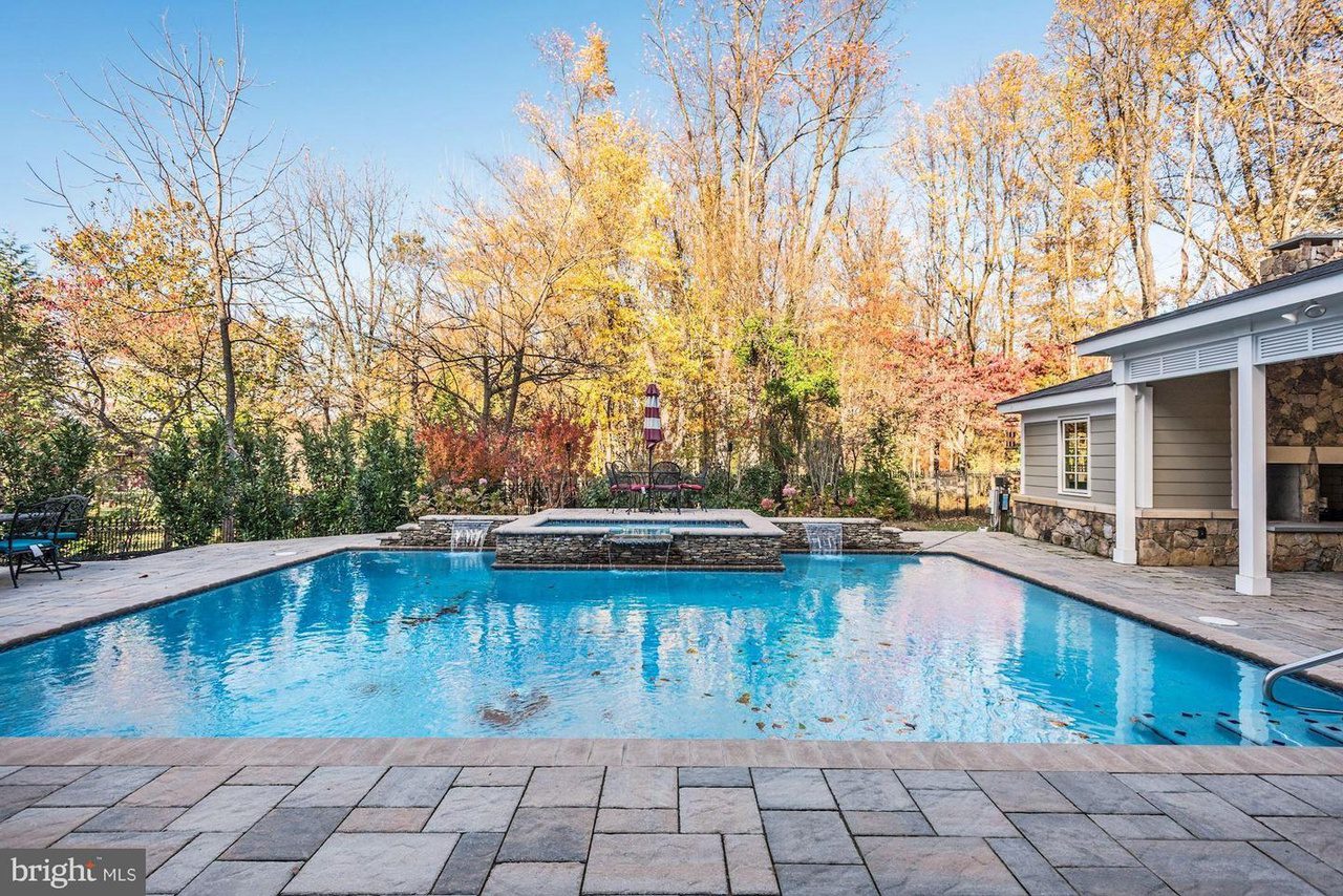 Outdoor pool with a stone patio, surrounded by trees with autumn foliage. A modern pool house is on the right.